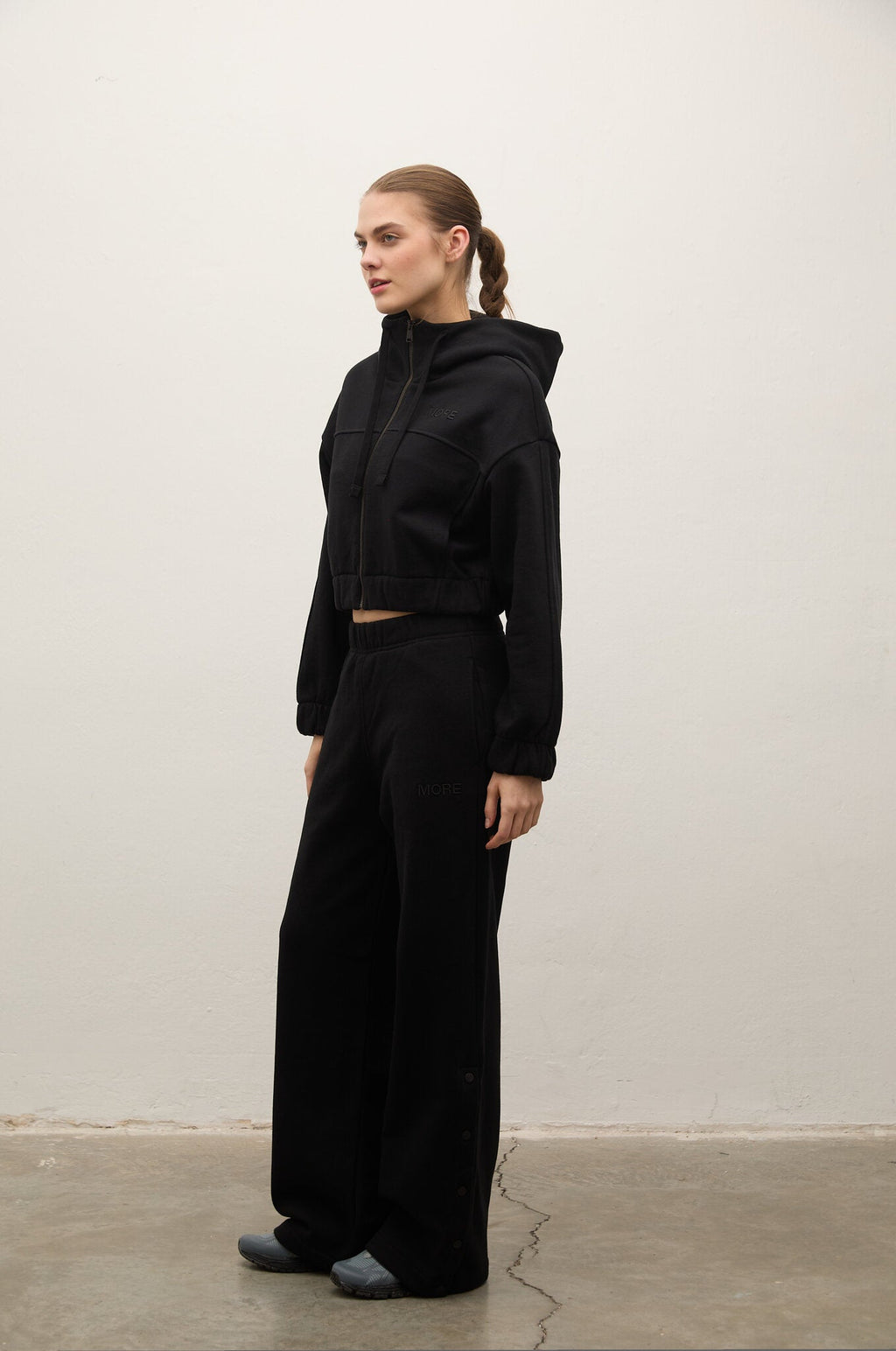 A woman stands in profile wearing a black cropped zip-up hoodie and matching black wide-leg sweatpants against a plain off-white wall and concrete floor.