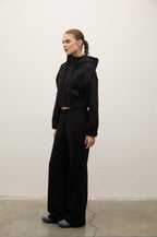 A woman stands in profile wearing a black cropped zip-up hoodie and matching black wide-leg sweatpants against a plain off-white wall and concrete floor.