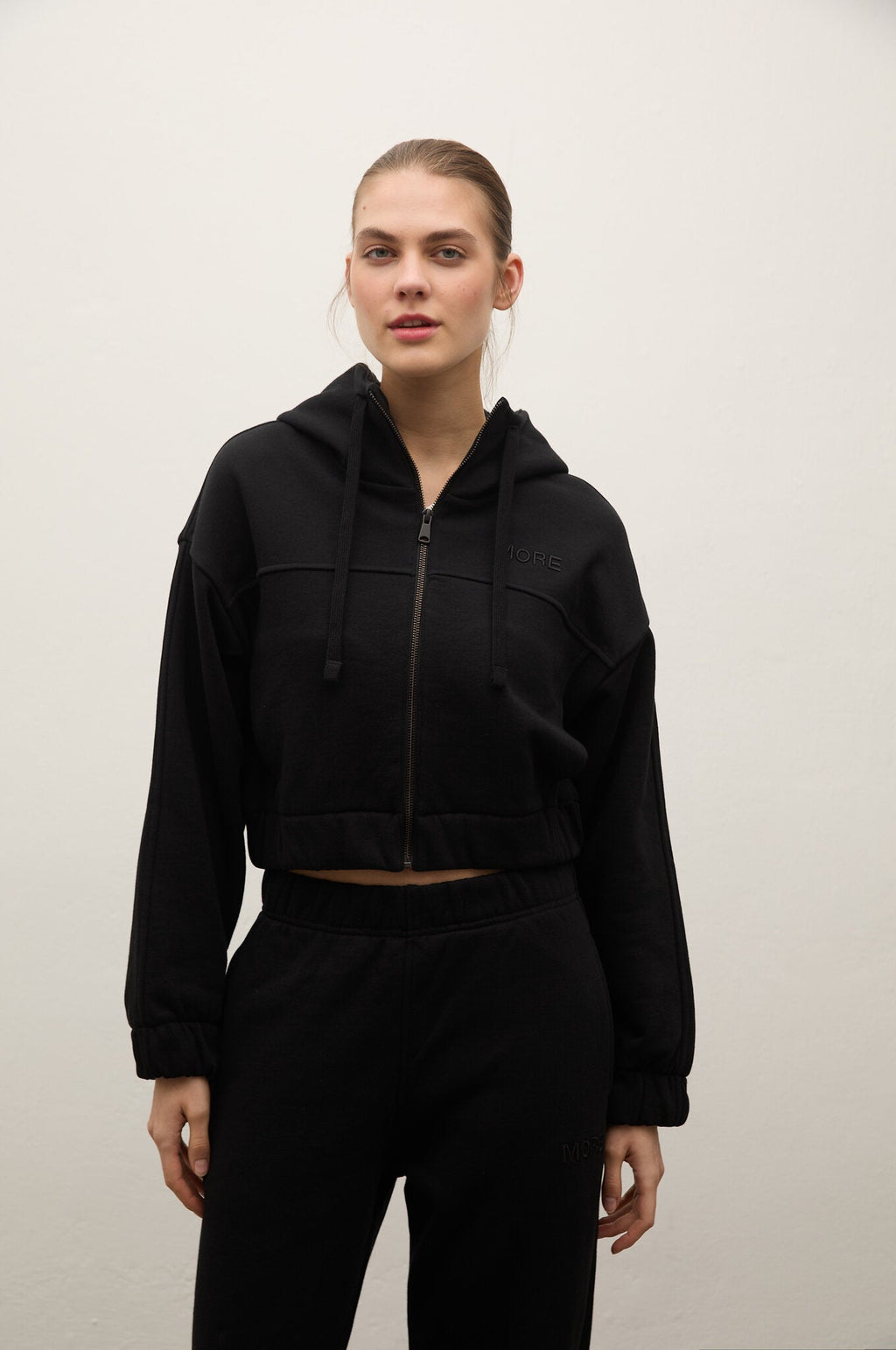 A woman modeling a black cropped zip-up hoodie with drawstrings and matching black sweatpants against a plain off-white wall.