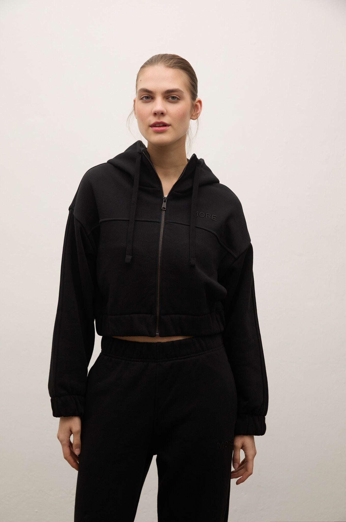 A woman modeling a black cropped zip-up hoodie with drawstrings and matching black sweatpants against a plain off-white wall.
