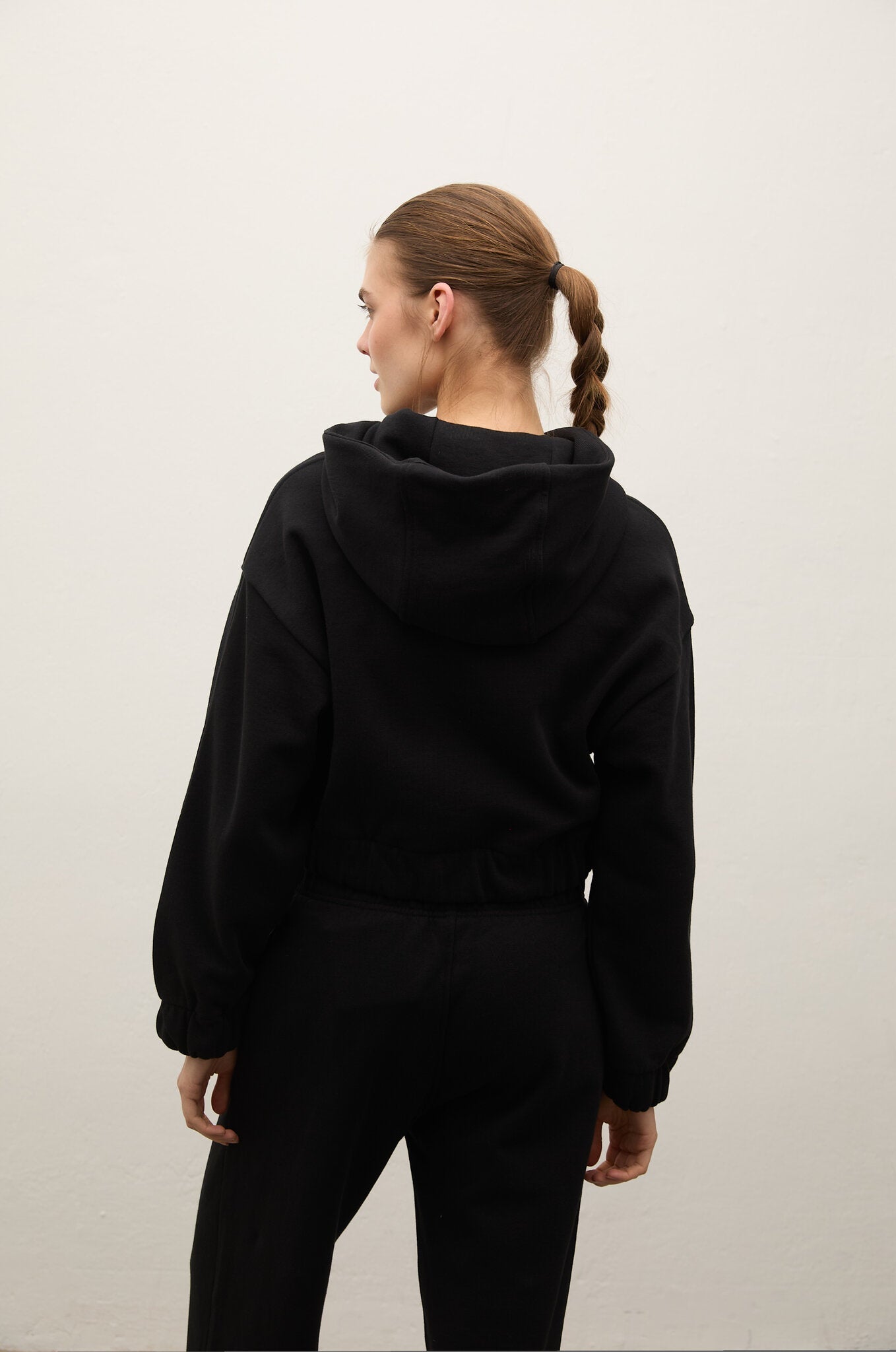 A woman with her hair in a single braid is shown from behind wearing a black cropped hoodie and matching black sweatpants against a plain off-white background.