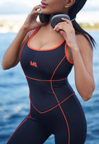 A woman wearing a black one-piece Gym Unitard with bright orange trim and orange piping details, standing outdoors with water in the background and holding a phone.