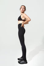 A woman modeling black athletic wear, featuring a black sports bra with a red and white logo band and matching black high-waisted leggings, paired with black Adidas sneakers, against a plain light gray studio background.