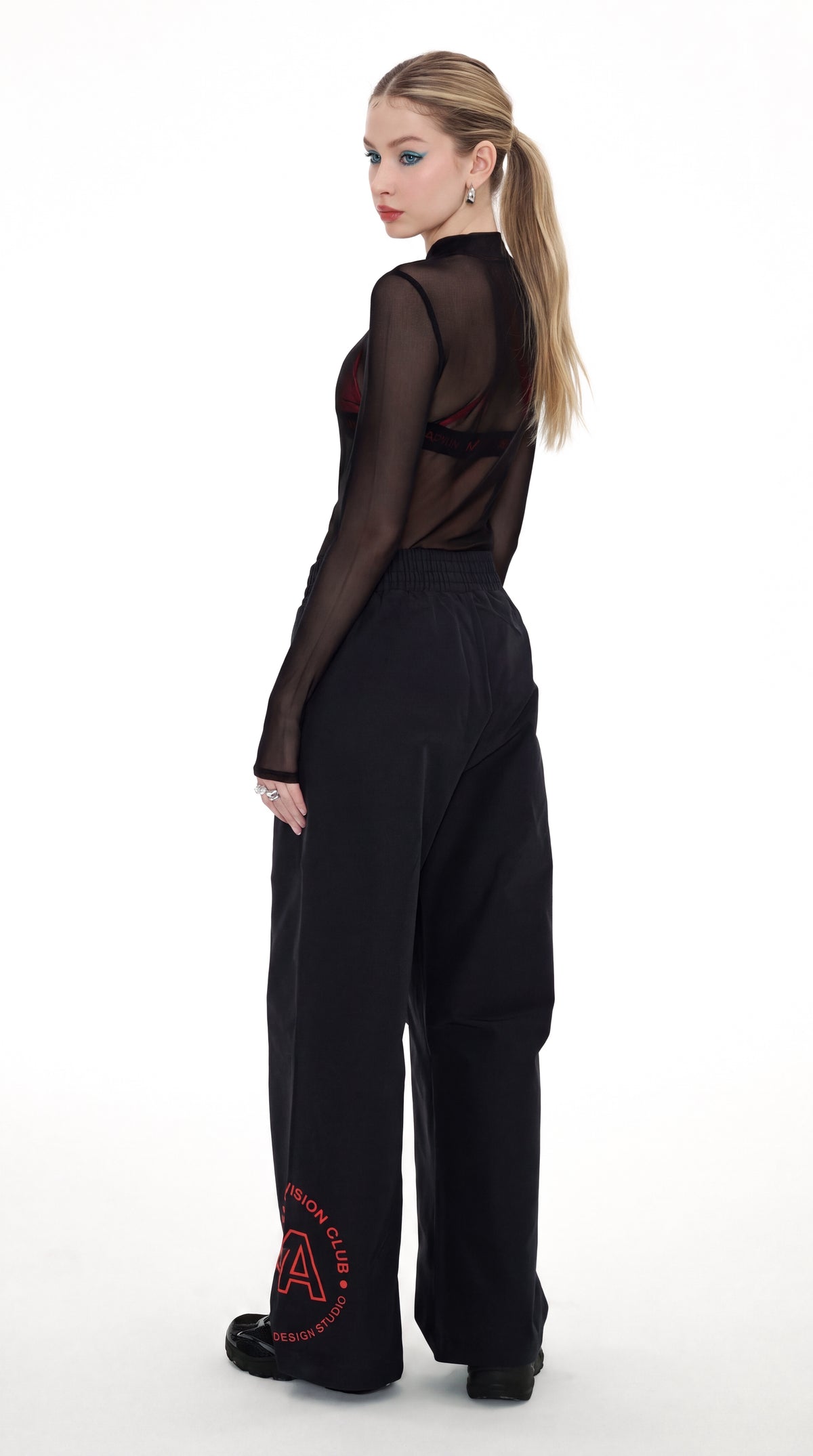 A woman stands in profile against a white studio background, wearing a sheer black long-sleeve top with a cutout back and black wide-leg pants featuring a red circular graphic near the hem.