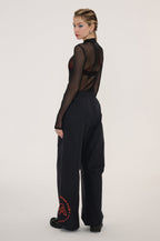 A model is shown from the back wearing a sheer black long-sleeve top with a high neck and wide-leg black trousers featuring a red circular graphic on the left leg, set against a plain light gray studio background.