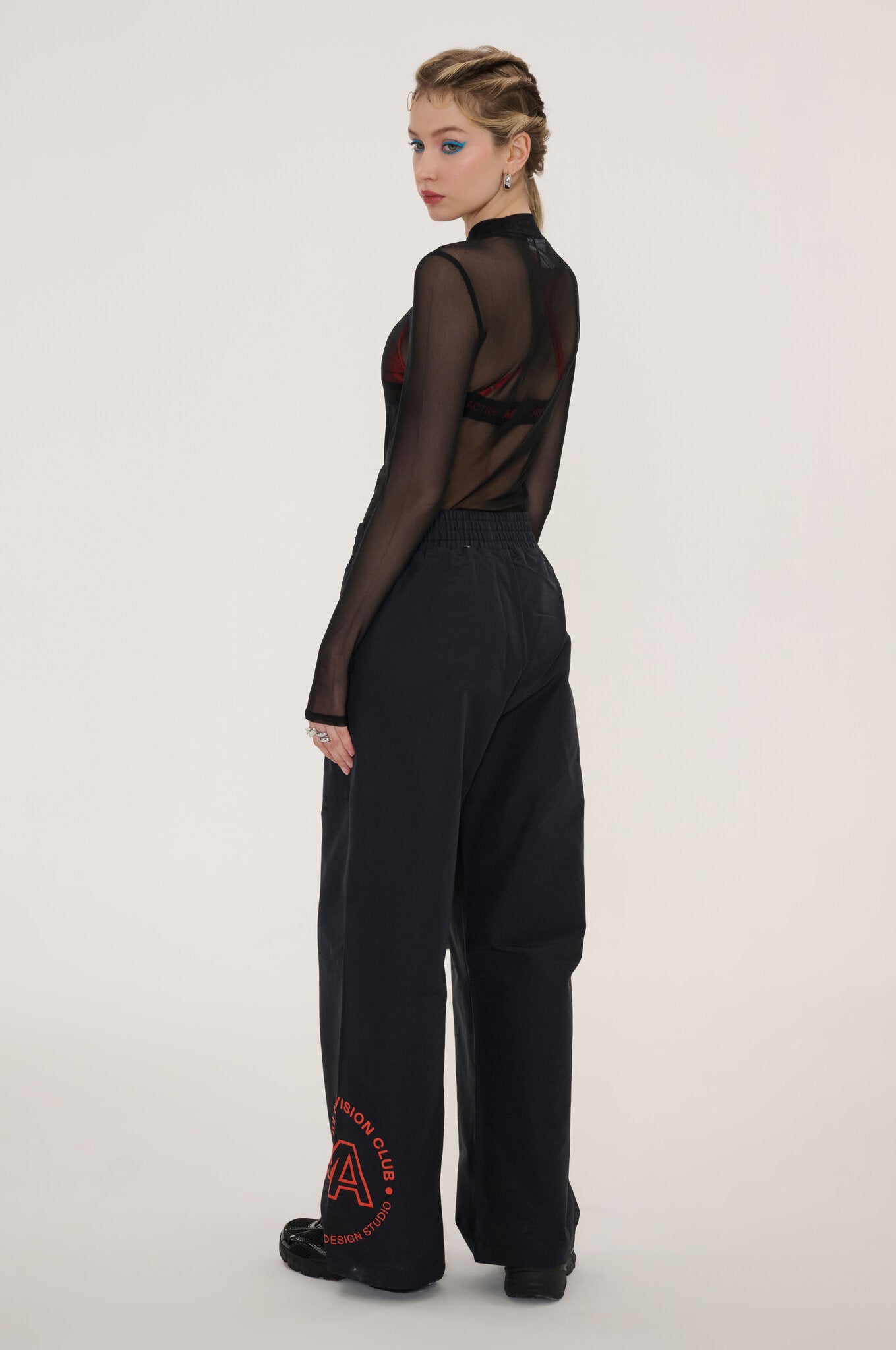 A model is shown from the back wearing a sheer black long-sleeve top with a high neck and wide-leg black trousers featuring a red circular graphic on the left leg, set against a plain light gray studio background.