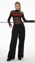 A model stands against a white studio background wearing a sheer black long-sleeve top with a red underlayer and black wide-leg pants with red graphic details.