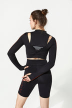 A woman wearing a matching black activewear set, featuring a long-sleeve crop top with cutout back details and black biker shorts, shown from a rear three-quarter view against a plain white studio background.