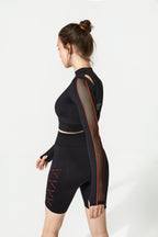 A woman modeling a black athletic set consisting of a long-sleeve mock-neck crop top with sheer mesh sleeves and matching high-waisted biker shorts, shown from a side profile against a light gray studio background.