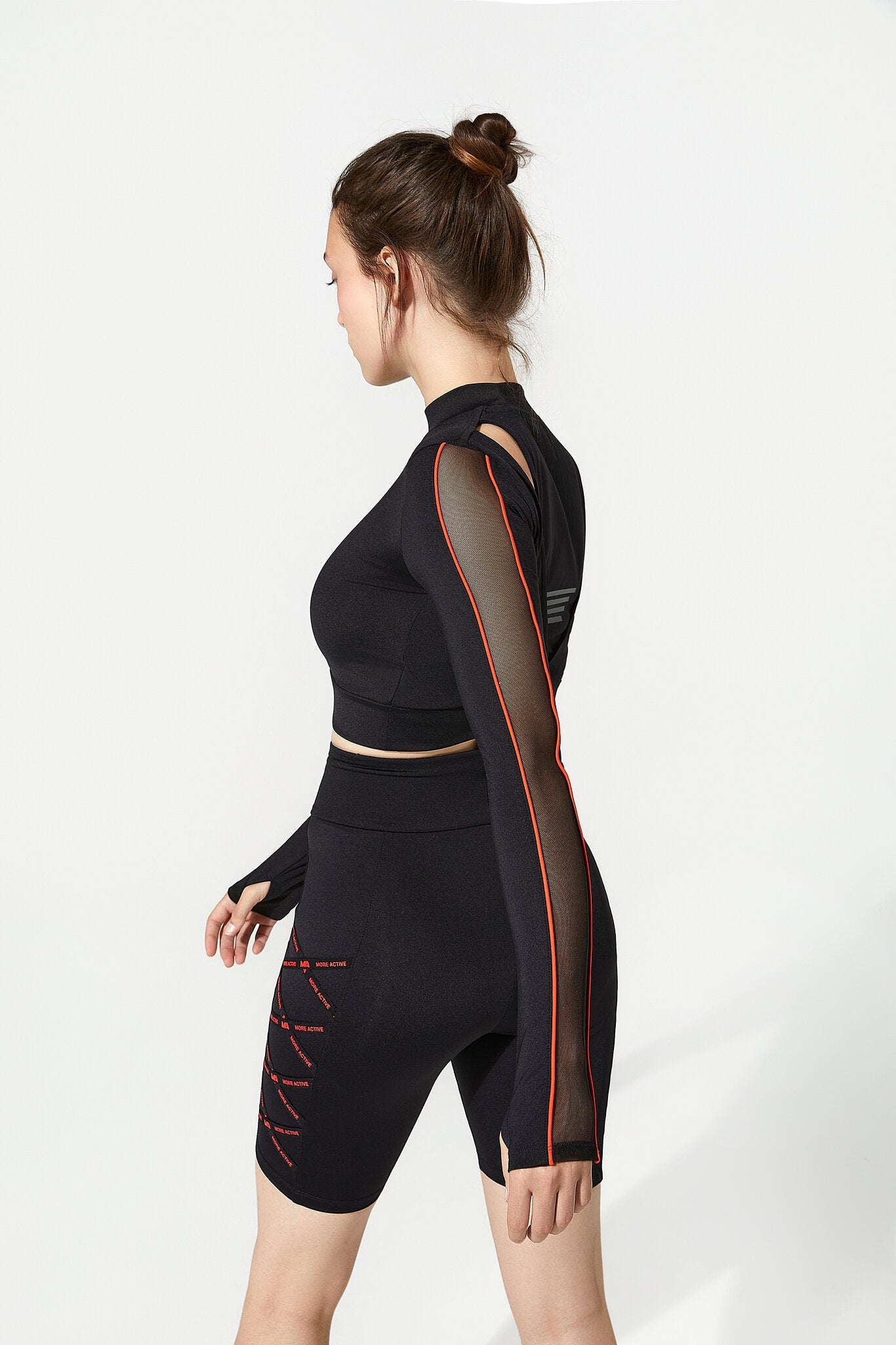 A woman modeling a black athletic set consisting of a long-sleeve mock-neck crop top with sheer mesh sleeves and matching high-waisted biker shorts, shown from a side profile against a light gray studio background.