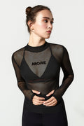 A woman modeling a black sheer mesh long-sleeve top with a V-neckline and the word 'MORE' printed across the chest, set against a plain light gray background.