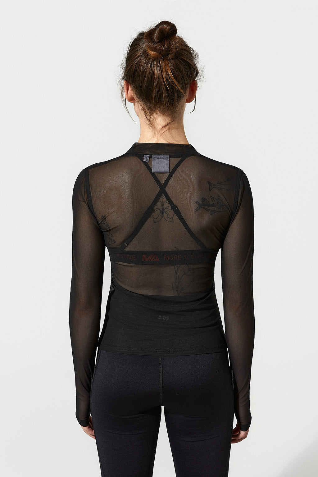 A woman viewed from the back wearing a black long-sleeve mesh top with an open crisscross back design and black leggings, against a plain light gray studio background.