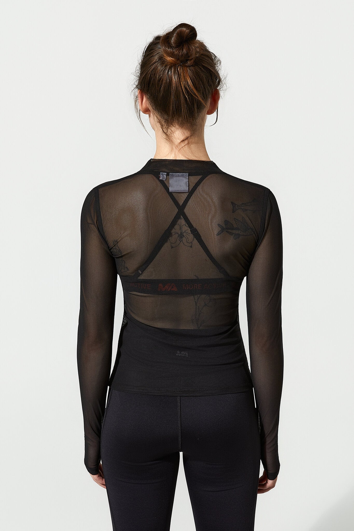 A woman viewed from the back wearing a black long-sleeve mesh top with an open crisscross back design and black leggings, against a plain light gray studio background.