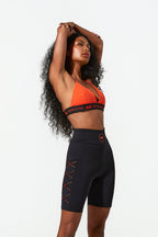 Woman wearing an orange sports bra and black shorts with red accents on a white background