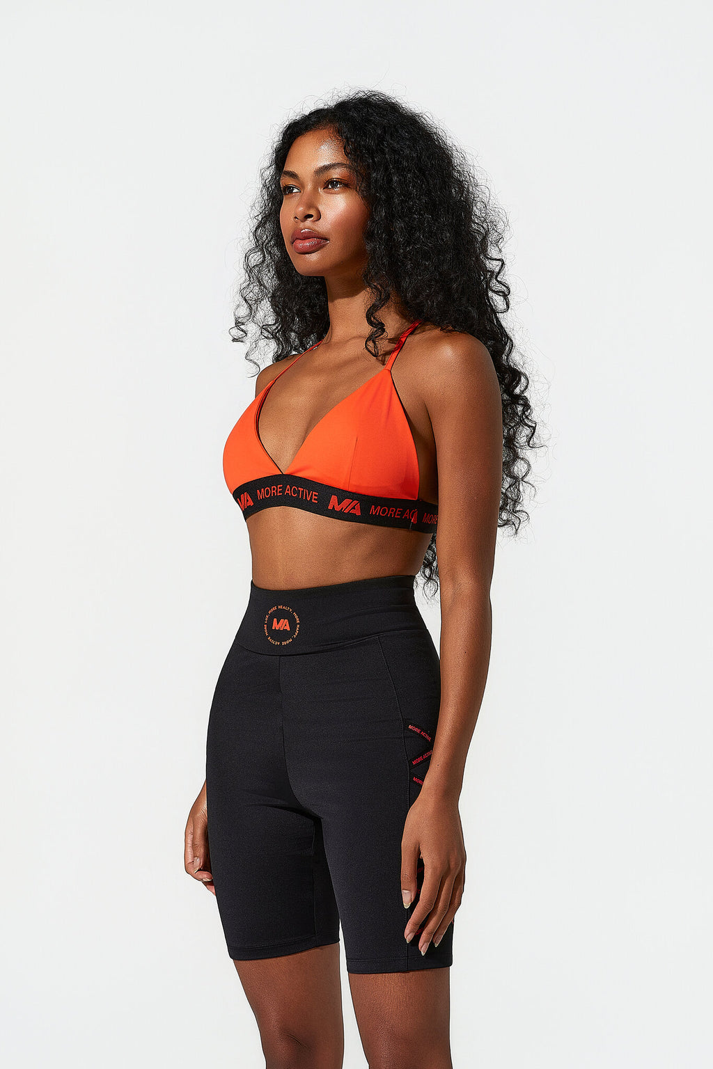 A woman modeling a matching activewear set consisting of an orange sports bra with black trim and black high-waisted biker shorts, photographed against a solid light gray background.