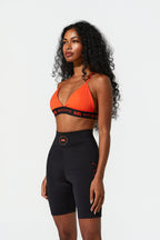 A woman modeling a matching activewear set consisting of an orange sports bra with black trim and black high-waisted biker shorts, photographed against a solid light gray background.