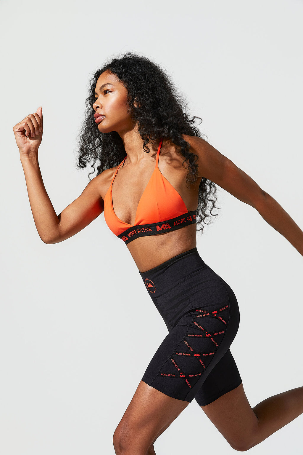 A woman in athletic wear posing dynamically against a light gray studio background. She is wearing a bright orange sports bra with black trim and matching black high-waisted shorts featuring red lace-up details on the side.