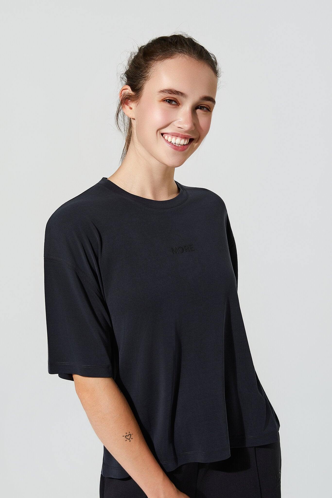 A woman smiling and wearing a loose-fitting, dark navy blue short-sleeve t-shirt with a small tonal logo on the left chest, set against a plain light gray studio background.