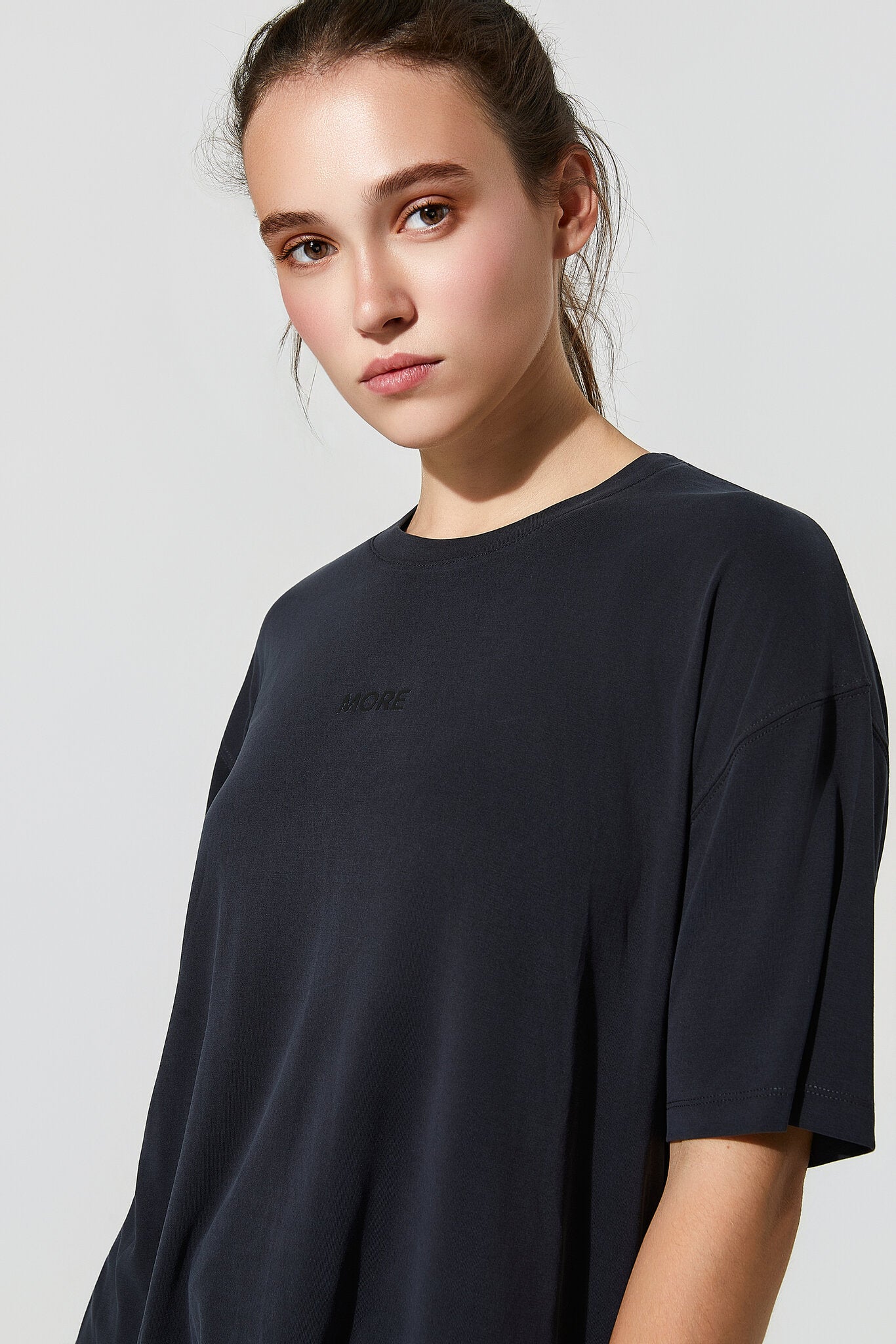 A woman wearing an oversized dark navy crewneck t-shirt with a subtle tonal logo on the left chest, photographed against a light gray studio background.