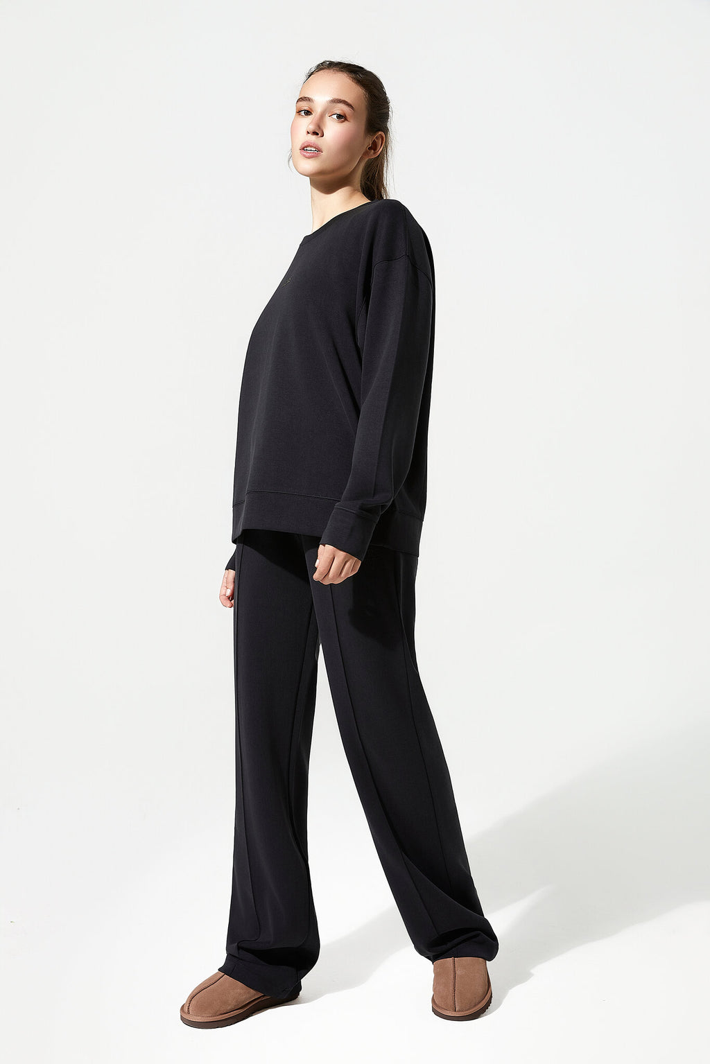 A woman modeling a black long-sleeve crewneck sweatshirt and matching black wide-leg sweatpants, paired with brown slip-on shoes, against a plain white studio background.