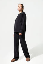 A woman modeling a black long-sleeve crewneck sweatshirt and matching black wide-leg sweatpants, paired with brown slip-on shoes, against a plain white studio background.