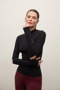 A woman modeling a black long-sleeve mock neck athletic top with built-in elbow sleeves, paired with burgundy leggings, against a plain light gray background.