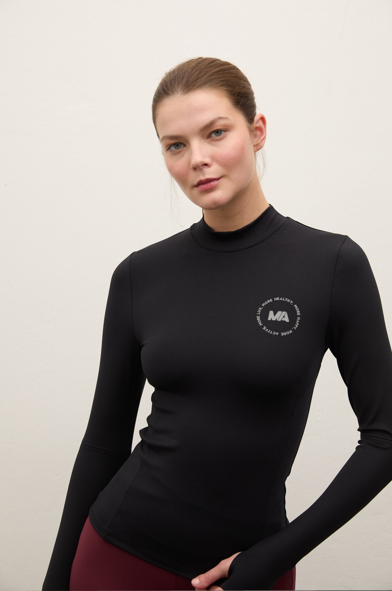A woman wearing a fitted black long-sleeve mock neck top with a small circular white logo on the left chest, paired with burgundy leggings, against a plain light background.