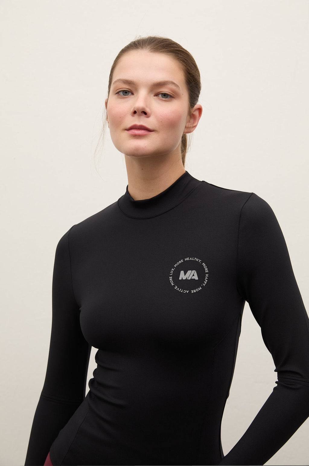 A woman wearing a black long-sleeve mock neck athletic top with a small white circular 'MA' logo on the left chest, photographed against a plain light gray studio background.