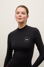 A woman wearing a black long-sleeve mock neck athletic top with a small white circular 'MA' logo on the left chest, photographed against a plain light gray studio background.