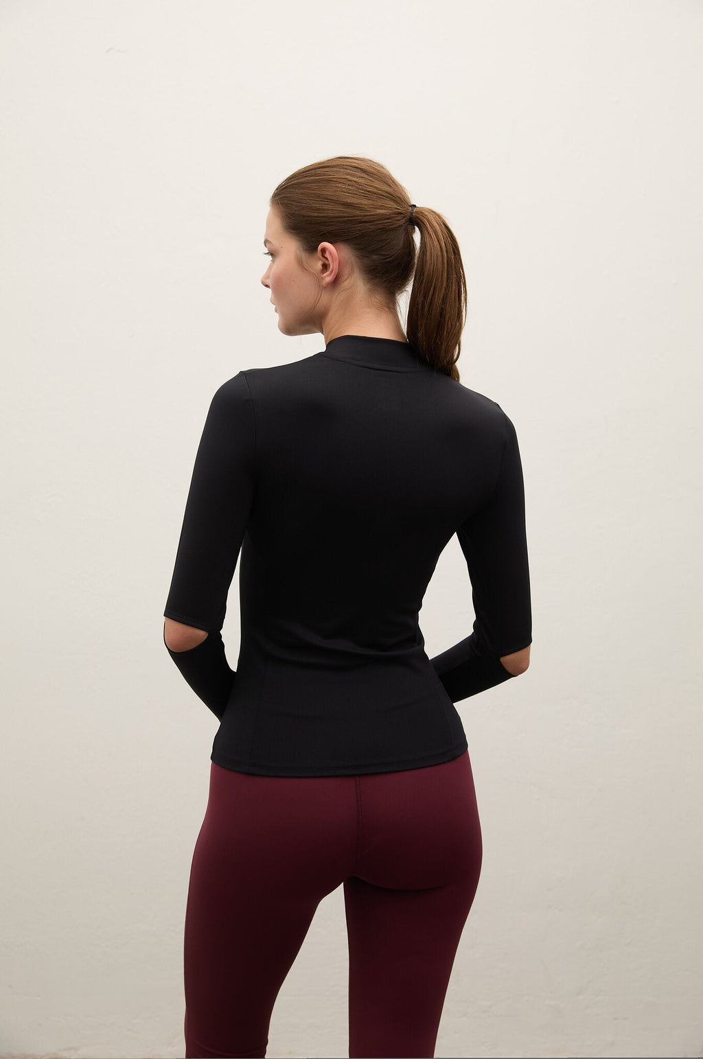 A woman shown from the back wearing a fitted black long-sleeve mock-neck athletic top and burgundy leggings, standing against a plain off-white wall.