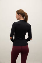 A woman shown from the back wearing a fitted black long-sleeve mock-neck athletic top and burgundy leggings, standing against a plain off-white wall.