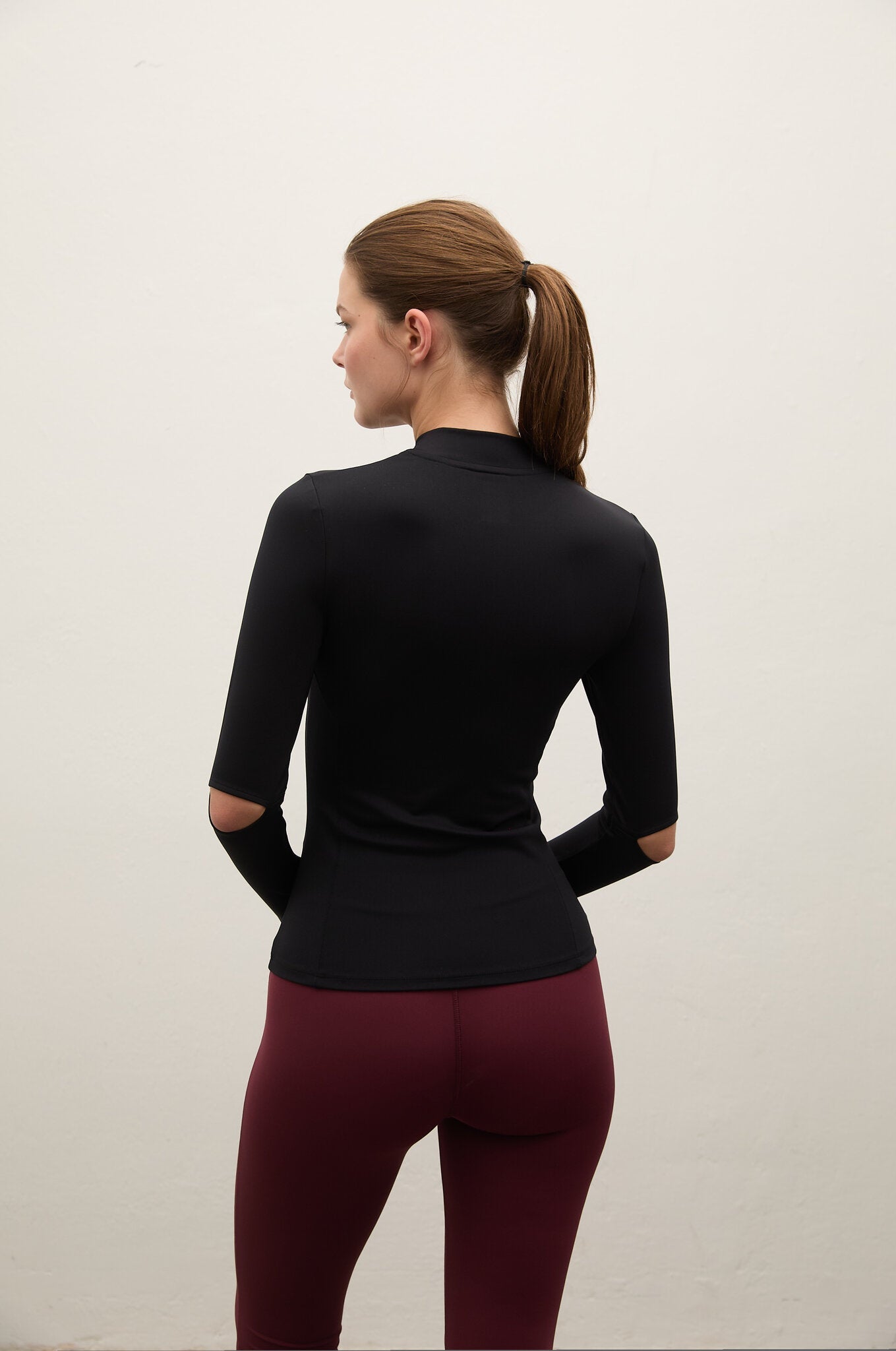 A woman shown from the back wearing a fitted black long-sleeve mock-neck athletic top and burgundy leggings, standing against a plain off-white wall.