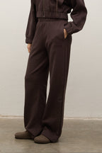 A person wearing dark brown wide-leg pants with side buttons and a matching cropped top, standing against a plain light wall.
