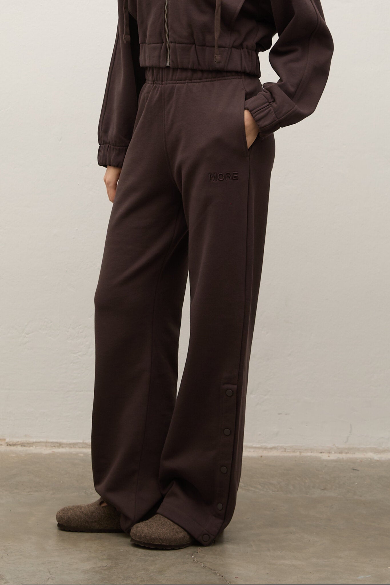 A person wearing dark brown wide-leg pants with side buttons and a matching cropped top, standing against a plain light wall.