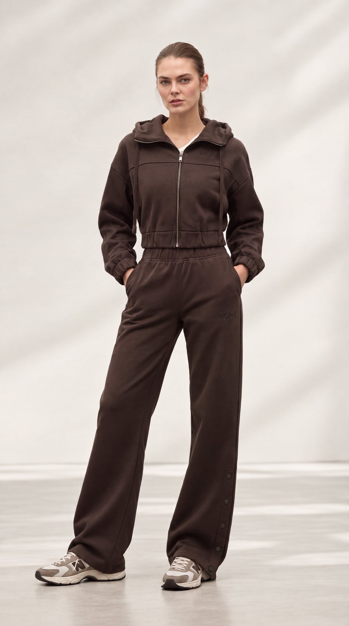 A woman modeling a dark brown zip-up hooded sweatshirt and matching wide-leg sweatpants, standing in a studio with a light gray backdrop.