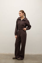 A woman modeling a dark brown zip-up hooded jumpsuit with wide-leg pants, standing in profile against a plain off-white wall.