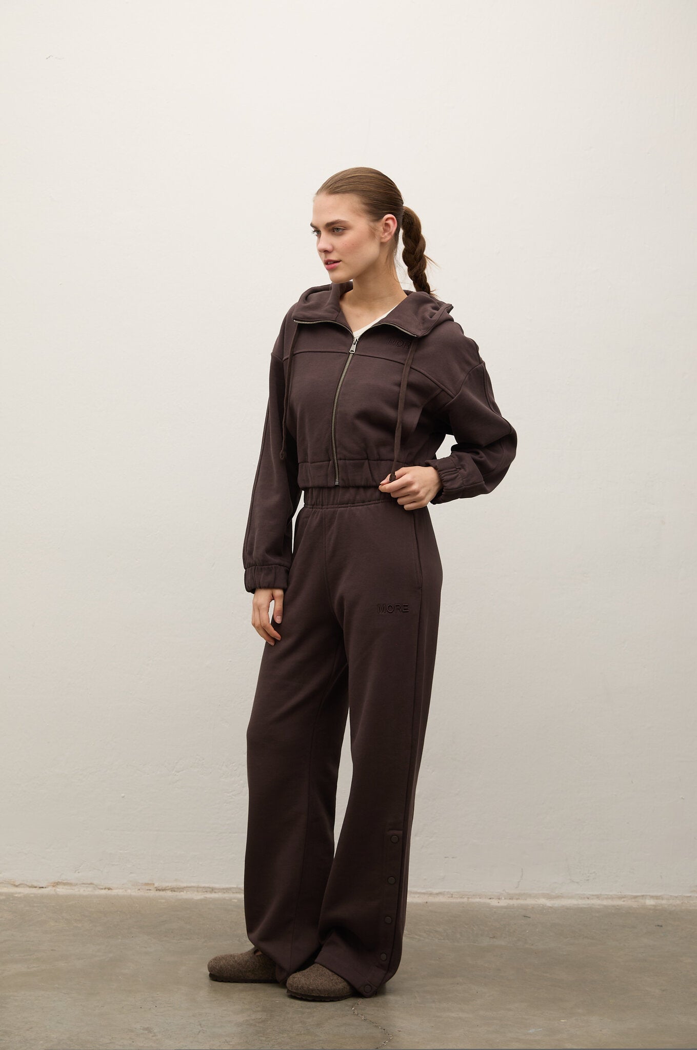 A woman modeling a dark brown zip-up hooded jumpsuit with wide-leg pants, standing in profile against a plain off-white wall.