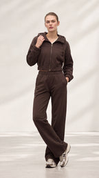 A model wearing a dark brown two-piece tracksuit consisting of a cropped zip-up jacket and matching wide-leg sweatpants, paired with gray and white sneakers, set against a minimalist light gray studio backdrop.