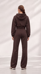 A woman viewed from behind wearing a matching dark brown cropped hoodie and wide-leg sweatpants set, standing against a light gray studio backdrop with soft diagonal shadows.