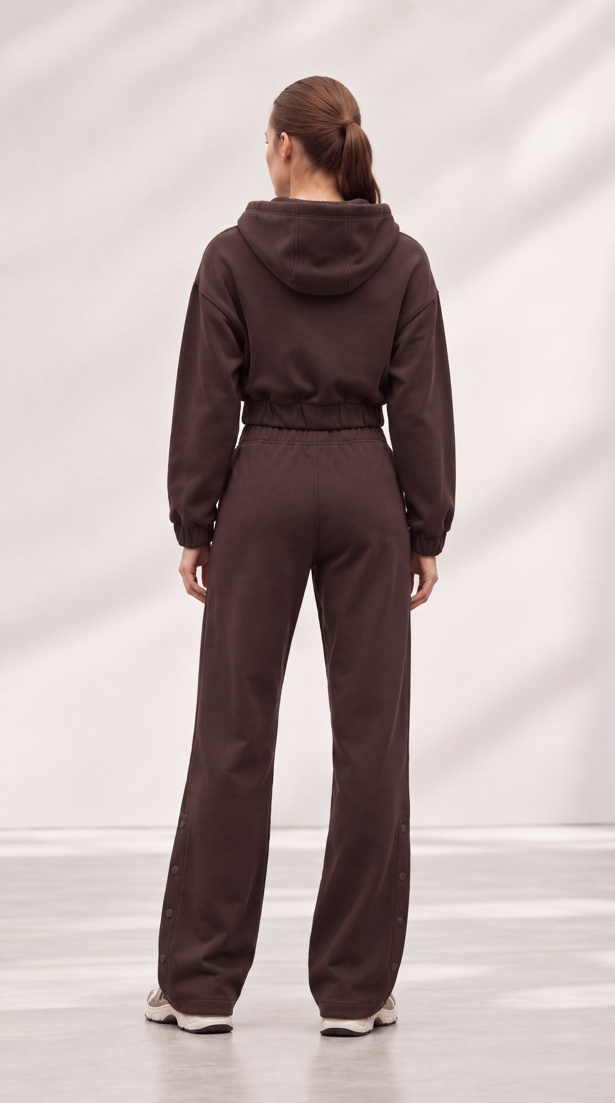 A woman viewed from behind wearing a matching dark brown cropped hoodie and wide-leg sweatpants set, standing against a light gray studio backdrop with soft diagonal shadows.