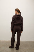 A woman stands with her back to the camera wearing a matching dark brown hooded sweatshirt and wide-leg sweatpants set, photographed in a minimalist studio with a white wall and concrete floor.