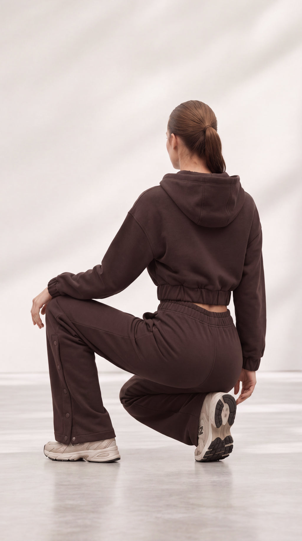 A woman wearing a matching dark brown cropped hoodie and sweatpants set, photographed from behind while crouching in a studio with a light gray background.