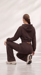 A woman wearing a matching dark brown cropped hoodie and sweatpants set, photographed from behind while crouching in a studio with a light gray background.