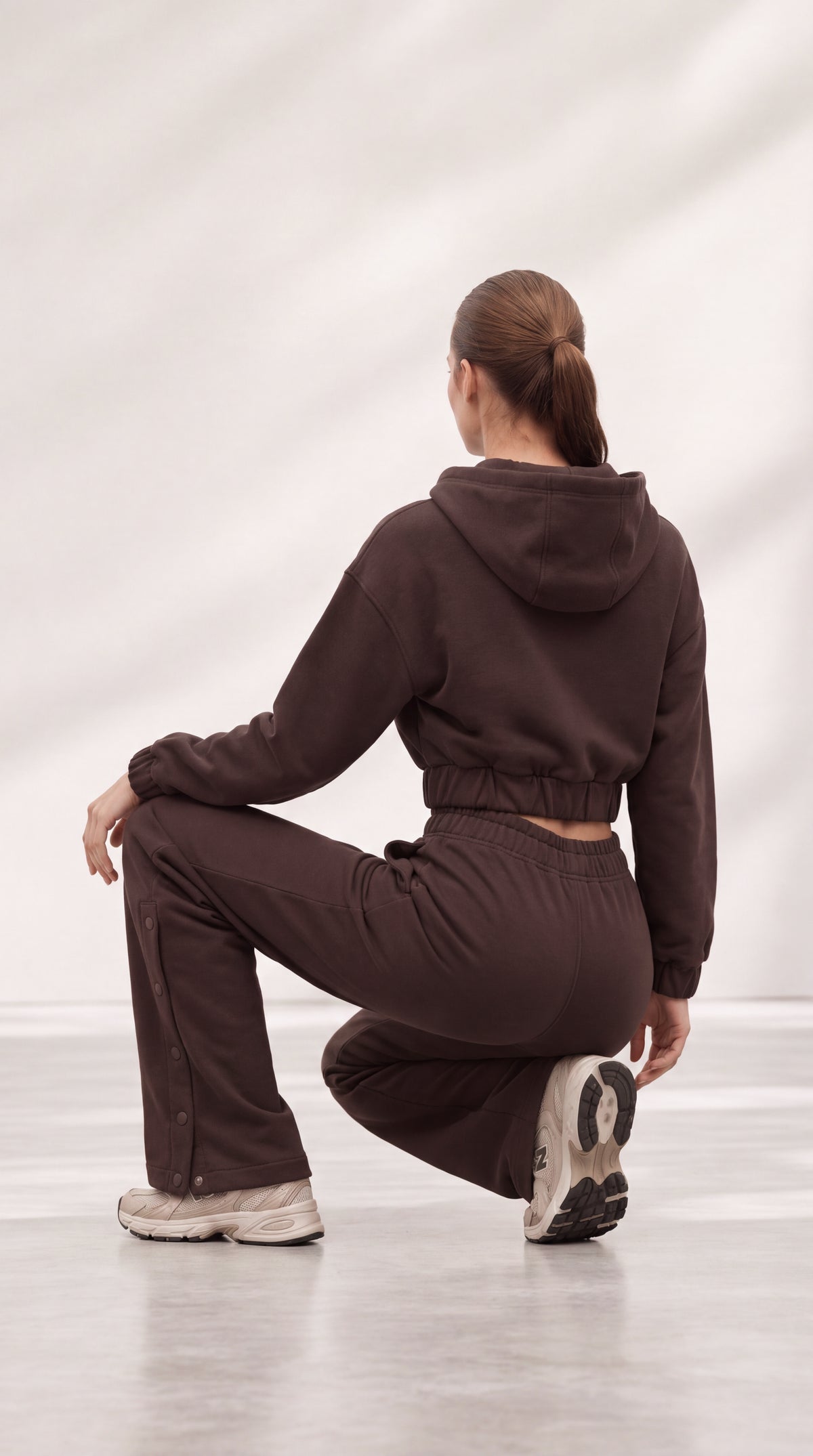 A woman wearing a matching dark brown cropped hoodie and sweatpants set, photographed from behind while crouching in a studio with a light gray background.