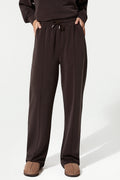 A person wearing dark brown wide-leg sweatpants with an elastic drawstring waist, paired with a matching dark top and brown slip-on shoes, photographed against a clean white studio background.