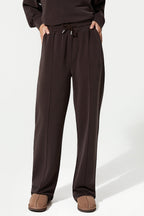 A person wearing dark brown wide-leg sweatpants with an elastic drawstring waist, paired with a matching dark top and brown slip-on shoes, photographed against a clean white studio background.
