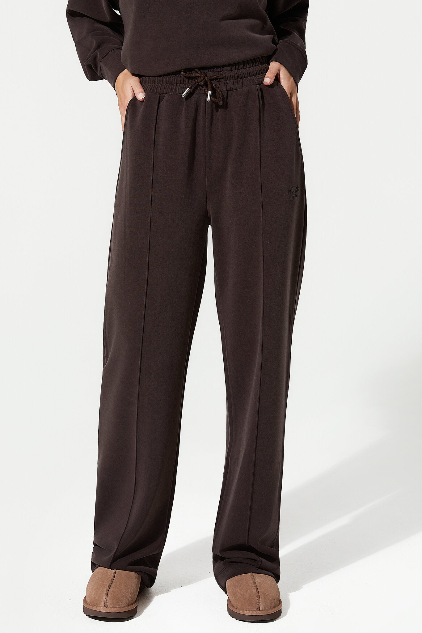 A person wearing dark brown wide-leg sweatpants with an elastic drawstring waist, paired with a matching dark top and brown slip-on shoes, photographed against a clean white studio background.