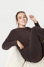 Person wearing a dark brown outfit sitting on a white surface with a plain background