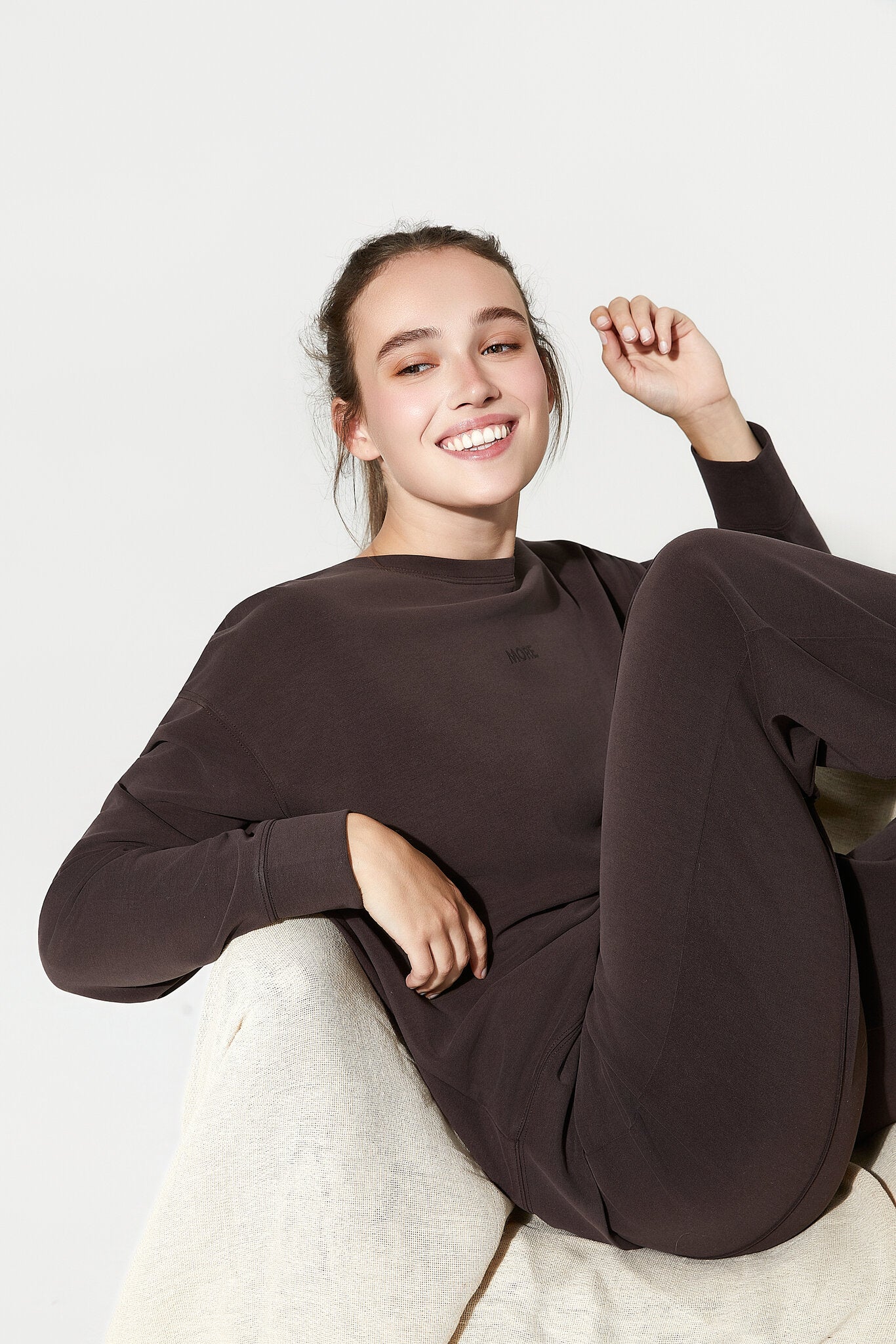 Person wearing a dark brown outfit sitting on a white surface with a plain background