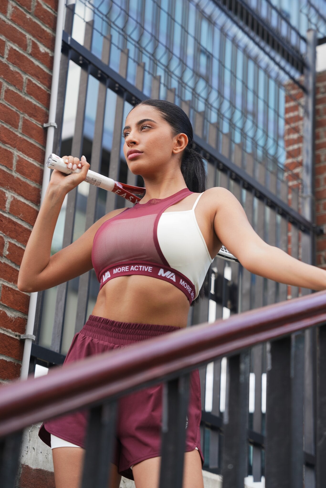 A woman wearing a matching athletic set consisting of a sports bra and shorts in maroon and white, holding a rolled-up towel over her shoulder while leaning on a black metal railing in front of a brick building.